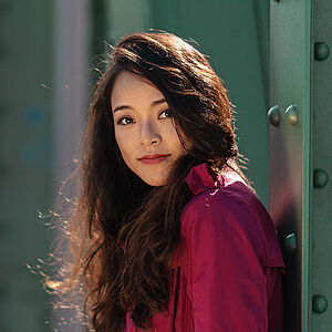 Portraitfoto von Ting-Yu Tsai. Sie trägt einen fuchsiafarbenen Mantel und ihr dunkles Haar offen. Sie lehnt an einer hohen grünen Stahlstrebe.