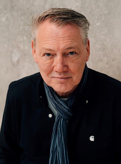 Portraitfoto von Dirk Böhling. Sein Haar ist grau und zur Seite gegelt. Er trägt eine dunkelblaue Jacke und einen in Blautönen gestreifter Schal. Er blickt von unten hinauf. Der Hintergrund ist beigefarben.