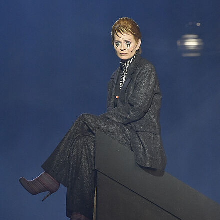 Eine Frau sitzt mit überschlagenen Beinen auf einer Erhöhung und schaut nach vorne. Ihre Augen sind schwarz unterlaufen.