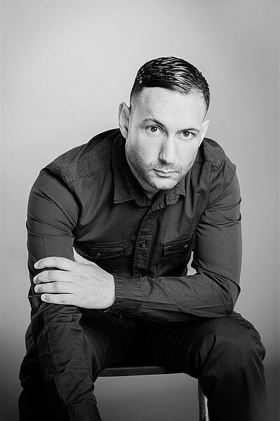 Schwarzweißes Portraitfoto von Barish Karademir. Er sitzt auf einem Stuhl und lehnt nach vorn. Der rechte Arm lehnt an seinem Knie und hängt herab. Der linke Arm liegt auf dem linken Knie, seine Hand greift den rechten Arm. Er trägt dunkles zurückgegeltes Haar und einen Dreitagebart. Er trägt ein dunkles Hemd und eine dunkle Jeans.