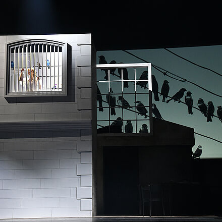 Links im Foto ist ein helles Gebäude mit einem hoch liegenden, großen Gitterfenster. Eine Frau in einem weißen Kleid steht am Gitterfenster und schaut nach draußen. Unten rechts im Foto steht ein Mann mit einem großen Rucksack. Er schaut nach oben, in Richtung Gitterfenster.