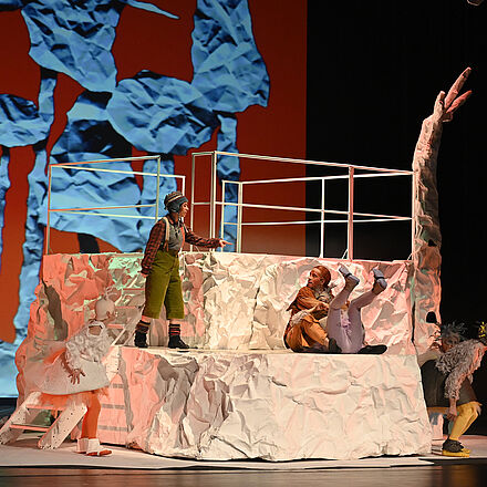 Auf dem Podest steht eine kleine Person, sie zeigt auf den Fuchs, der eine Wildgans im Arm hält. Unten stehen zwei weitere Gänse und verstecken sich. Im Hintergrund erkennt man einen Wald aus Papier