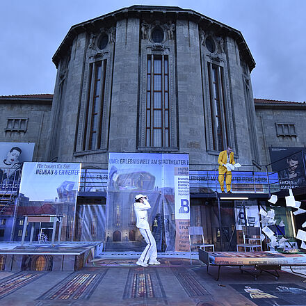 Man erkennt eine Bühne vor dem Stadttheater. Es fliegen Zettel.