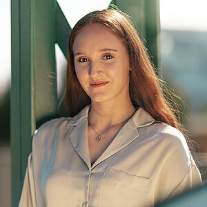 Portraitfoto von Melissa Festa. Sie trägt ihr rotes langes Haar offen und grinst. Sie lehnt an einem grünen Stahlgerüst. Sie trägt eine beige Bluse, dazu eine Kette mit goldenem Anhänger.