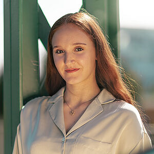 Portraitfoto von Melissa Festa. Sie trägt ihr rotes langes Haar offen und grinst. Sie lehnt an einem grünen Stahlgerüst. Sie trägt eine beige Bluse, dazu eine Kette mit goldenem Anhänger.