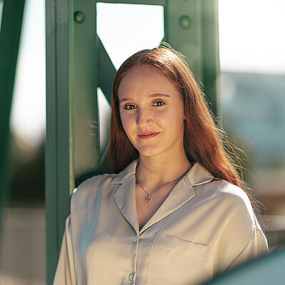 Portraitfoto von Melissa Festa. Sie trägt ihr rotes langes Haar offen und grinst. Sie lehnt an einem grünen Stahlgerüst. Sie trägt eine beige Bluse, dazu eine Kette mit goldenem Anhänger.