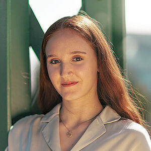 Portraitfoto von Melissa Festa. Sie trägt ihr rotes langes Haar offen und grinst. Sie lehnt an einem grünen Stahlgerüst. Sie trägt eine beige Bluse, dazu eine Kette mit goldenem Anhänger.
