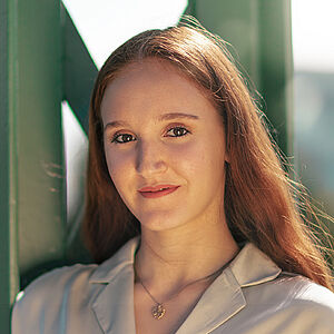 Portraitfoto von Melissa Festa. Sie trägt ihr rotes langes Haar offen und grinst. Sie lehnt an einem grünen Stahlgerüst. Sie trägt eine beige Bluse, dazu eine Kette mit goldenem Anhänger.