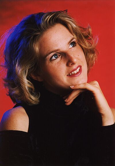 Portraitfoto von Yvonne Blunk. Sie sitzt seitlich und blickt nach links oben. Ihr Kinn ist auf ihrem linken Handrücken aufgestützt. Sie ist in schwarz gekleidet. Ihr Schultern liegen frei. Ihr blondes kinnnlanges Haar wellt sich. Der Hintergrund ist rot.