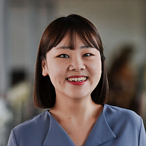 Portraitfoto von Minji Kim. Sie lächelt mit offenem Mund. Sie trägt schulterlanges dunkelbraunes Haar und einen Pony, dazu roten Lippenstift. Sie ist in eine fliederfarbene Bluse gekleidet. Der Hintergrund ist verschwommen. 