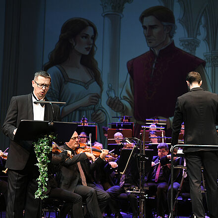 Im Vordergrund steht eine Person, das Orchester spielt, und im Hintergrund ist ein Bild einer Frau und eines Mannes zu sehen.  