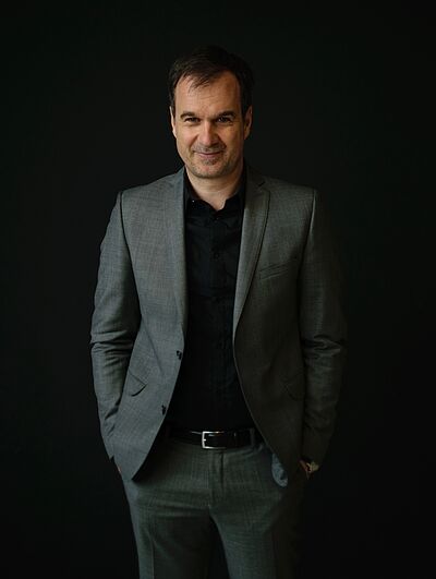 Portraitfoto von Andreas Kloos. Er steht lächelnd vor einer dunklen Wand. Er hat braunes kurzes Haar. Er trägt einen grauen Anzug und ein schwarzes Hemd, dazu einen schwarzen Gürtel und eine Armbanduhr. Seine Hände sind in den Hosentaschen.