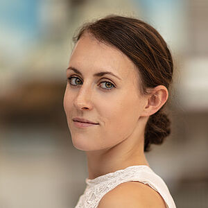 Portraitfoto von Annika Ellen Osenberg. Sie steht seitlich und schaut über die linke Schulter. Sie trägt ein weißes kurzärmliges Oberteil. Ihr Sie trägt ihr braunes Haar zu einem tiefen Dutt gebunden.