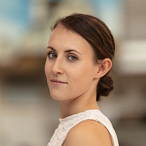 Portraitfoto von Annika Ellen Osenberg. Sie steht seitlich und schaut über die linke Schulter. Sie trägt ein weißes kurzärmliges Oberteil. Ihr Sie trägt ihr braunes Haar zu einem tiefen Dutt gebunden.