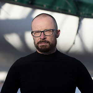 Portraitfoto von Krisztian Jambor. Er trägt einen schwarzen Rollkragenpullover und eine schwarze Brille. Er hat einen Bart und sehr kurze Haare.