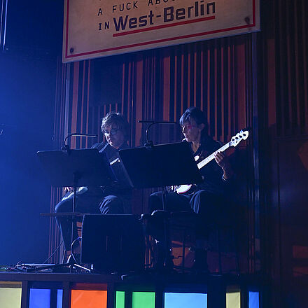 Auf einer bunten Erhöhung sitzen ein Gitarrist und eine Bassistin. Über den Musikern hängt ein Schild mit der Aufschrift «ACHTUNG They don't give a fuck about you in West-Berlin».