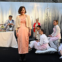 Im Vordergrund steht Roxane, grinsend zum Publikum gewandt. Sie trägt ein rosa Kleid und eine Wolljacke. Das Ensemble teils auf dem Boden sitzend, teils im Hintergrund, schaut Roxane an. 