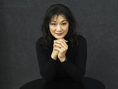 Portraitfoto von Mari Kodama. Sie sitzt an mit den Ellbogen auf einem schwarzen Tisch aufgelehnt. Sie trägt einen schwarzen Pullover. Ihr schwarzes Haar ist leicht gewellt und reicht bis auf die Schultern. Der Hintergrund ist dunkelgrau.