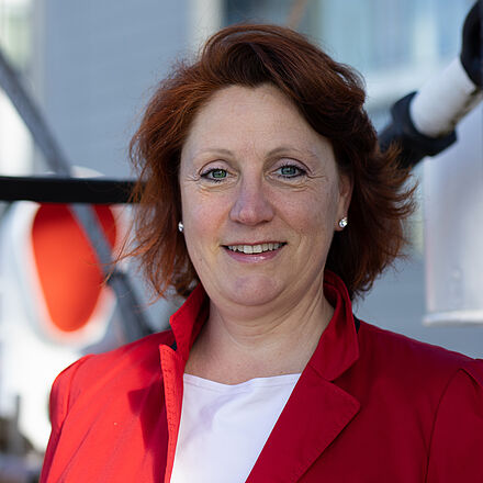 Portraitfoto von Diana Veiser. Sie blickt lächelnd in die Kamera. Sie trägt einen roten Blazer über einem weißen Oberteil. Sie hat rotbraunes Haar und trägt Perlenohrringe. 