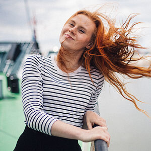 Portraitfoto von Marie-Theres Schwinn. Sie steht an der Reling eines Schiffes. Ihr rotes Haar weht im Wind. Sie trägt ein schwarz-weiß geringeltes Oberteil.