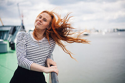 Portraitfoto von Marie-Theres Schwinn. Sie steht an der Reling eines Schiffes. Ihr rotes Haar weht im Wind. Sie trägt ein schwarz-weiß geringeltes Oberteil.