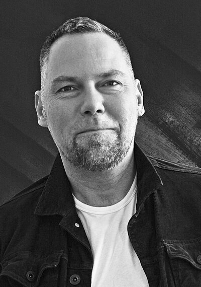 Schwarzweißes Portraitfoto von Walter Schütze. Er steht leicht schräg. Er trägt sein Haar kurz, dazu einen kurzen Bart um den Mund. Er trägt eine dunkles Jeansjacke zu einem weißen T-Shirt.