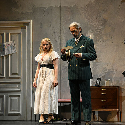 Ein Mann mit grauen Haaren steht in der Mitte des Bildes und blickt auf einen Brief in seiner Hand. Links hinter ihm steht eine Frau und schaut ihn an.