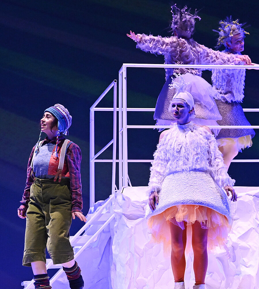 Eine kleine Person auf einem Podest aus Papier blickt in die Ferne; eine Gans steht neben ihr, während im Hintergrund Wildgänse tänzerisch durch die Luft ziehen.