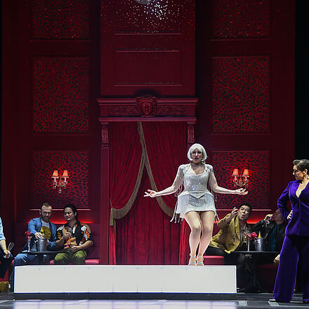 Im Hintergrund des Bildes sitzen mehrere Menschen in kleinen Sitzecken. Im Vordergrund steht eine Frau auf einem Podest und streckt die Arme zur Seite aus. Neben dem Podest stehen zwei Frauen mit Tablett und eine Frau in einem lila Anzug.