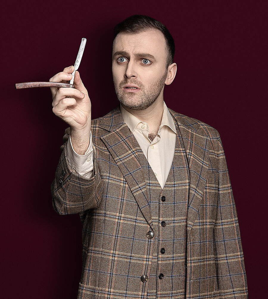Ein Mann (ANdrew Irwin) hält in seiner rechten Hand ein Rasiermesser. Als Kleidung trägt er einen karierten braunen Anzug und ein helles Hemd mit Weste drunter. Der Hintergrund des Bildes ist weinrot.
