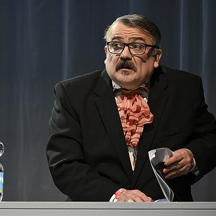 Ein Mann sitzt an einem Tisch, blättert in einem Heft und schaut mit großen Augen nach vorne.