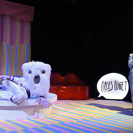 Auf einem Podest liegt eine Person. Links daneben steht eine Person am Mikro. Im Hintergrund steht «Alles Lüge?»