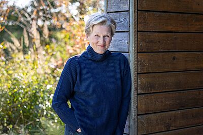 Portraitfoto von Angela Denoke. Sie trägt einen dunkelblauenn Rollkragenpullover und lehnt an einer Holzwand. Dahinter ist es begrünt. Ihre Hände stecken ind en Hosentaschen. Sie trägt ihr helles Haar kurz und lächelt verschmitzt.