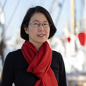 Portraitfoto von Tomoyo Ueda. Sie trägt einen schwarzen Pullover, dazu einen gebundenen roten Schal. Ihr dunkles Haar ist kinnlang. Sie trägt eine ovale Brille und lächelt dezent.