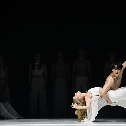 Im Hintergrund erkennt man Tänzer:innen. Sie beobachten einen Tänzer und eine Tänzerin. Die Tänzerin liegt auf den Knien des Tänzers.
