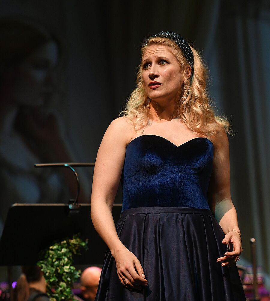 Im Vordergrund steht eine Frau in einem blauen Kleid, im Hintergrund befindet sich das Orchester.  