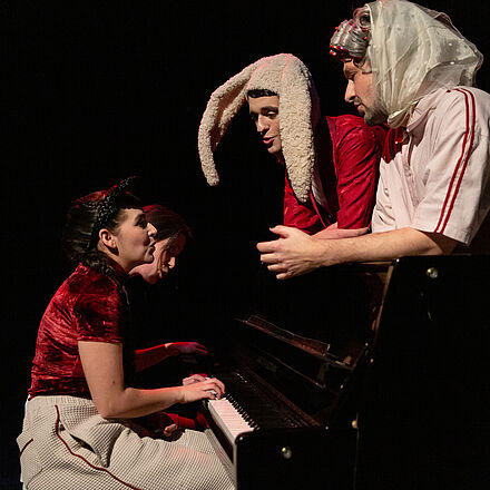 Zwei junge Frauen sitzen an einem Klavier und spielen gemeinsam. Zwei weitere Personen lehnen am Klavierkasten und singen gemeinsam mit den Spielenden.