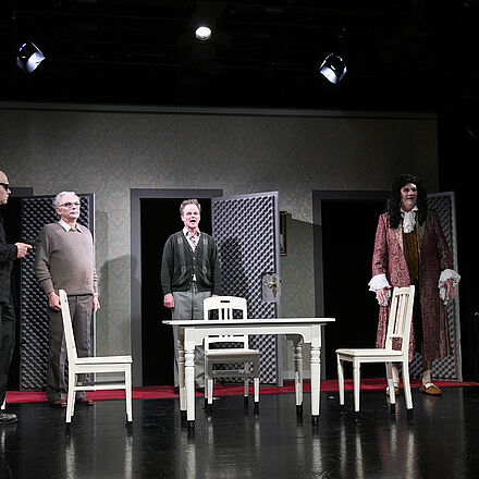 Im Mittelpunkt steht ein Tisch, um den mehrere Personen herumstehen. Rechts im Bild befindet sich ein zweiter Tisch, an dem eine Frau sitzt. Im Hintergrund sieht man drei Türen.