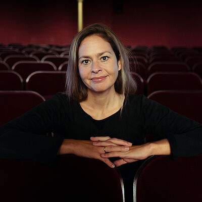 Portraitbild von Mascha Schubert. Sie sitzt in einem Theatersaal und hat die Hände verschrenkt. Sie trägt ein schwarzes T-Shirt und ihr dunkles Haar offen.