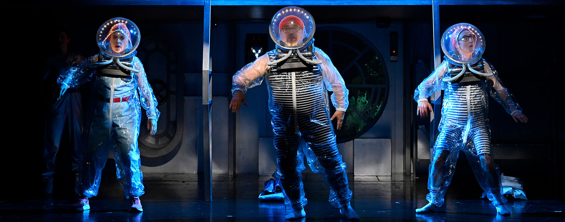 Drei Personen in durchsichtigen Taucheranzügen stehen auf der Bühne und schauen zum Publikum. Die Szene ist in blaues Licht getaucht.