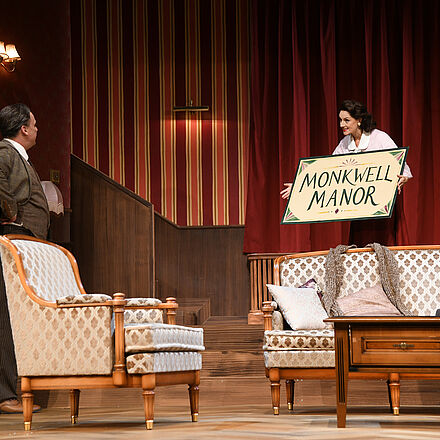 Der Fokus des Bildes liegt auf einer Frau, welche auf der rechten Seite eines älter anmutenden Salons ein Schild mit der Aufschrift «Monkwell Manor» hält. Sie beugt sich dabei mit dem Schild in Richtung eines Mannes, dessen Gesicht zu ihr gedreht und verdeckt ist. Im Vordergrund sieht man ein Sessel, sowie Sofa und Tisch.