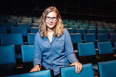 Portraitfoto von Christina Gegenbauer. Sie sitzt in einem Theatersaal voller blauer Klappsitze. Ihr Haar ist blond und lockig. Sie trägt ein Jeanshemd und eine Brille.