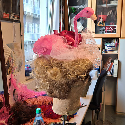 Der Hut ist auf einer Perücke mit blonden lockigen Haaren befestigt. Der Hut besteht aus einem pinken Flamingo und rosa Tüll.