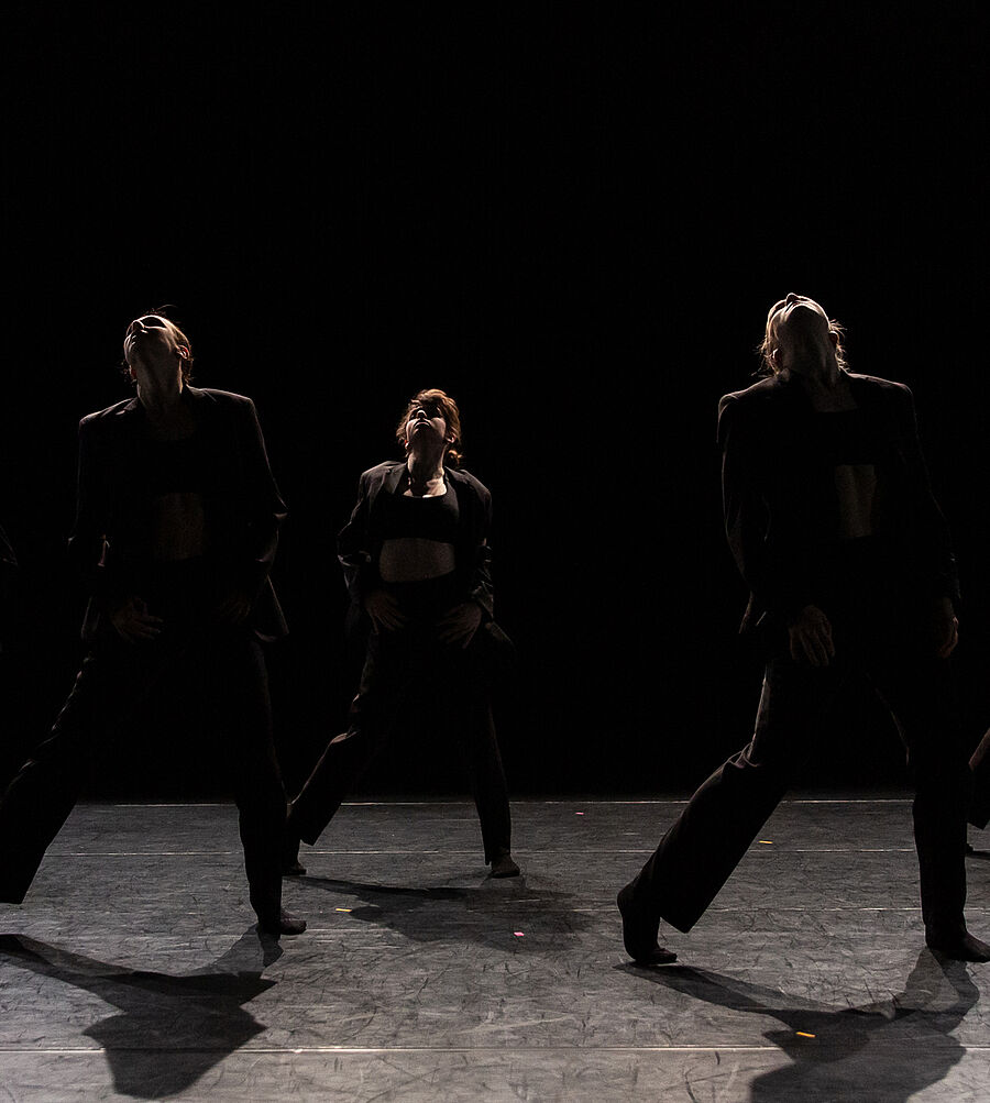 Fünf Tänzerinnen in zwei Reihen. Sie stehen in schummrigen Licht, die Bühne ist nahezu schwarz. Sie tragen schwarze Anzüge zu bauchfreien Tops und blicken nach oben. Ihre Beine sind gespreizt, ihre Hände liegen auf ihren Oberschenkeln. Die Scheinwerfer leuchten von der Decke.