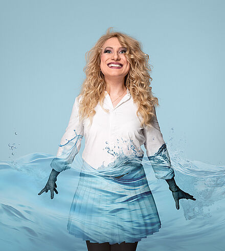 Eine Frau mit blonden Locken und weißer Bluse und Rock steht im Wasser. Ihre Hände sind im Wasser.