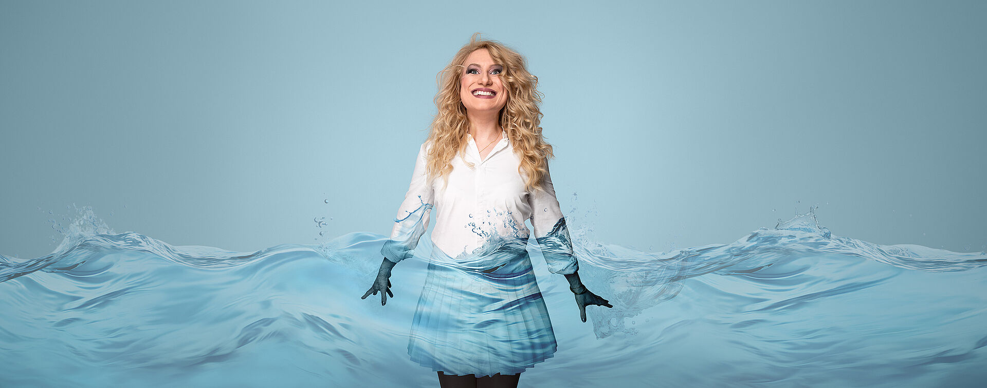 Eine Frau mit blonden Locken und weißer Bluse und Rock steht im Wasser. Ihre Hände sind im Wasser.