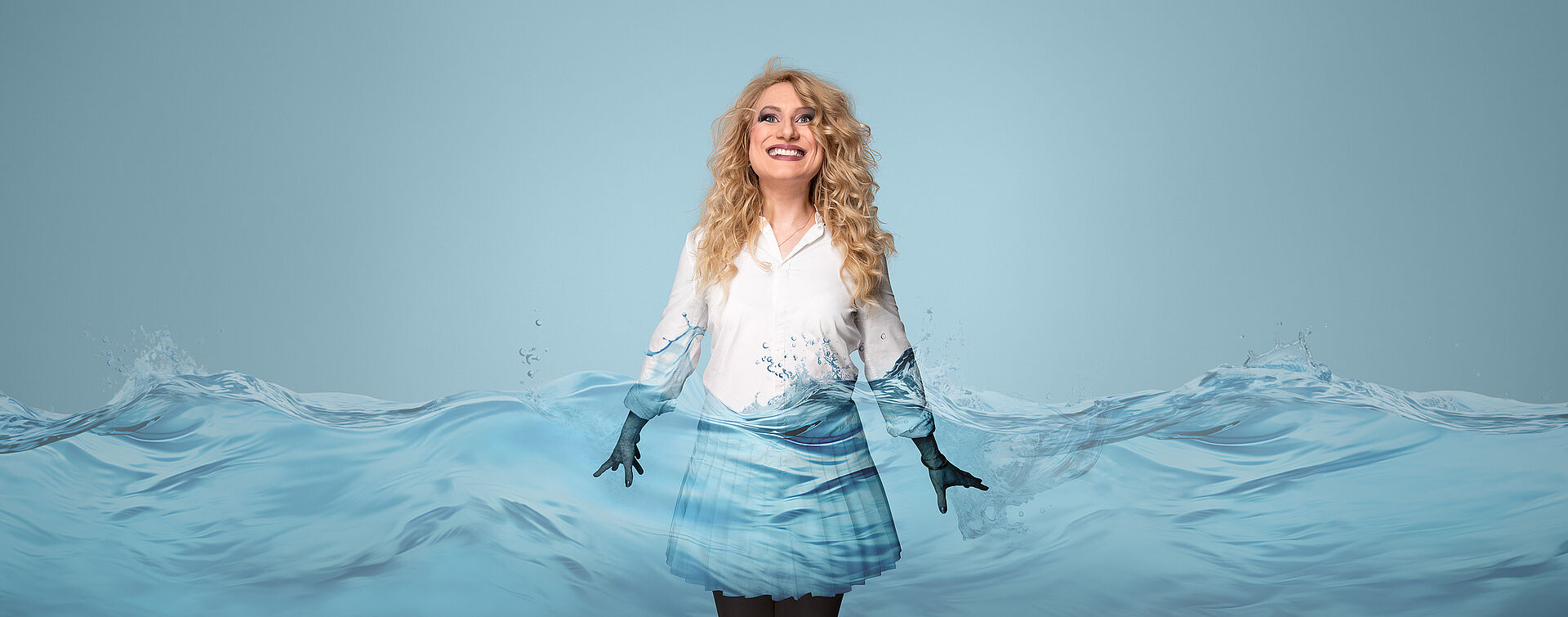 Eine Frau mit blonden Locken und weißer Bluse und Rock steht im Wasser. Ihre Hände sind im Wasser.