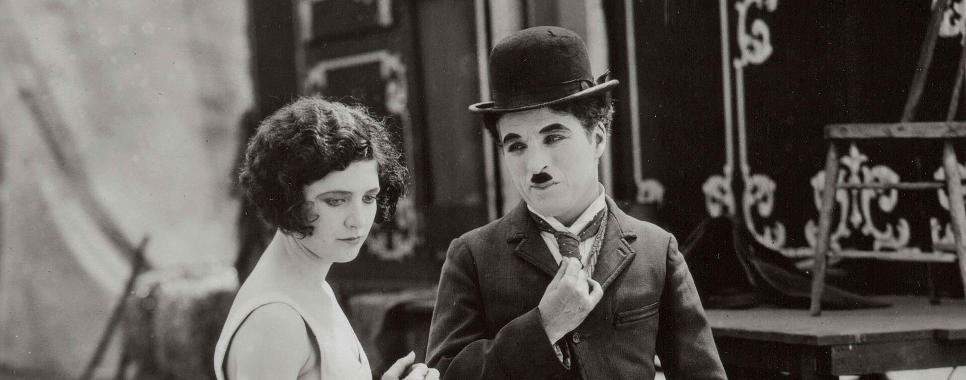 Charlie Chaplin blickt auf eine Frau. Hinter ihnen stehen Zirkuswagen.