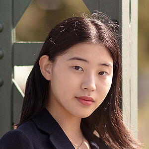 Portraitfoto von Dawon Yang. Sie trägt einen dunkelblauen Blazer über einem schwarzen Oberteil. Ihr schwarzes Haar fällt ihr über die rechte Schulter. Sie lehnt an einem grünen Stahlgerüst.