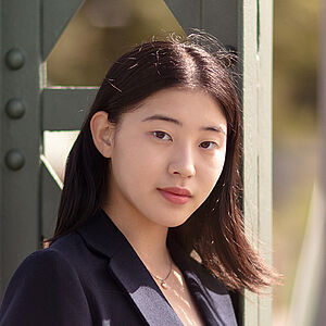 Portraitfoto von Dawon Yang. Sie trägt einen dunkelblauen Blazer über einem schwarzen Oberteil. Ihr schwarzes Haar fällt ihr über die rechte Schulter. Sie lehnt an einem grünen Stahlgerüst.